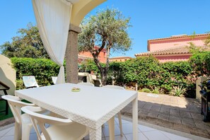 Une terrasse avec une table et des chaises blanches, entourée de verdure et bordée d’un bâtiment dont le toit est recouvert de tuiles.