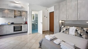 Ein modernes Hotelzimmer mit Küchenzeile, einem Bett und ordentlich gefalteten Handtüchern, sowie einem durchgehenden Türrahmen einsehbares Badezimmer.