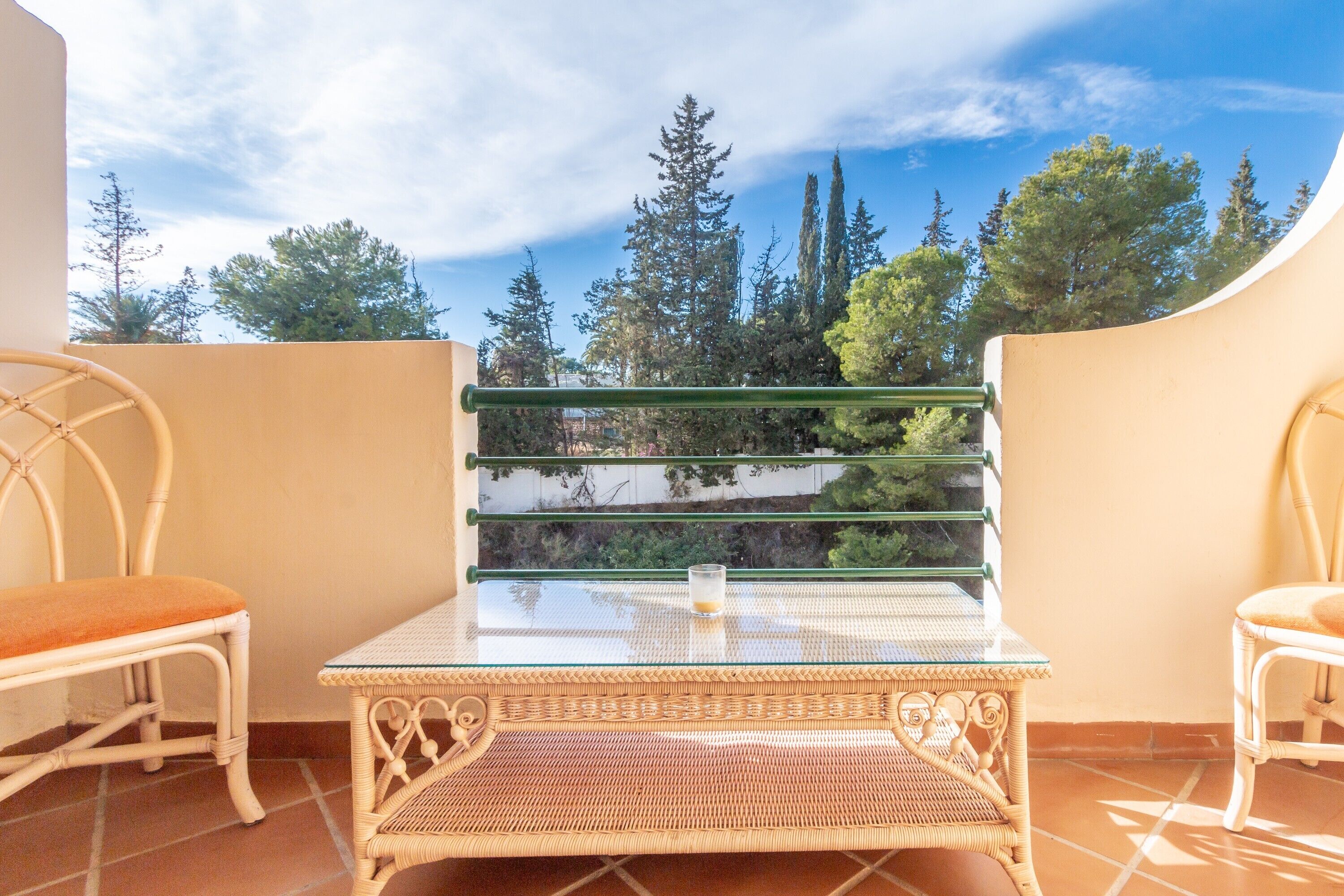 A balcony with wicker furniture, a glass table, and a view of trees and sky.