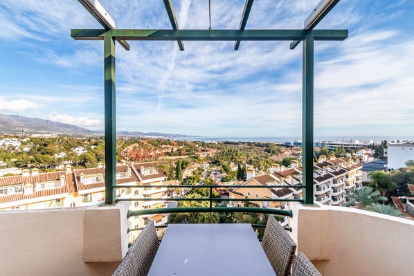 A balcony with a table and chairs, overlooking a coastal town with buildings and a body of water.