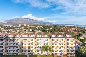 A multi-story residential building with balconies, a mountainous backdrop, and a clear sky.
