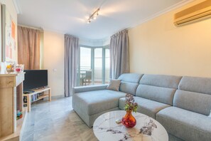 A modern living room with a grey sofa, a coffee table with a vase, a flat-screen TV, and a bookshelf.