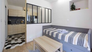 Une petite pièce comprenant un lit, une petite table, un coin cuisine et une étagère fixée au mur, sur laquelle est posé un sapin de Noël.