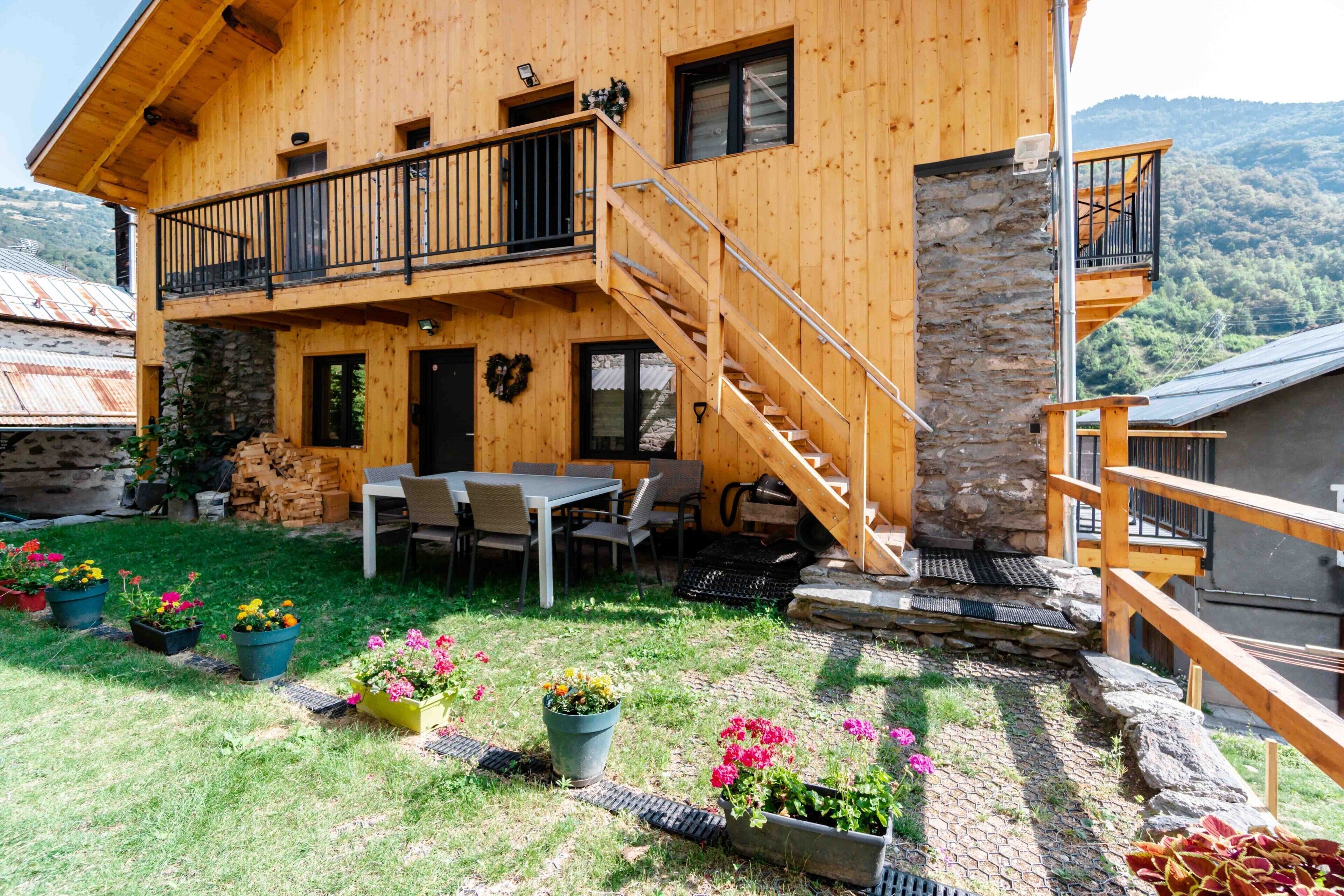 A wooden cabin with a stone wall, a balcony, a staircase, and outdoor seating.
