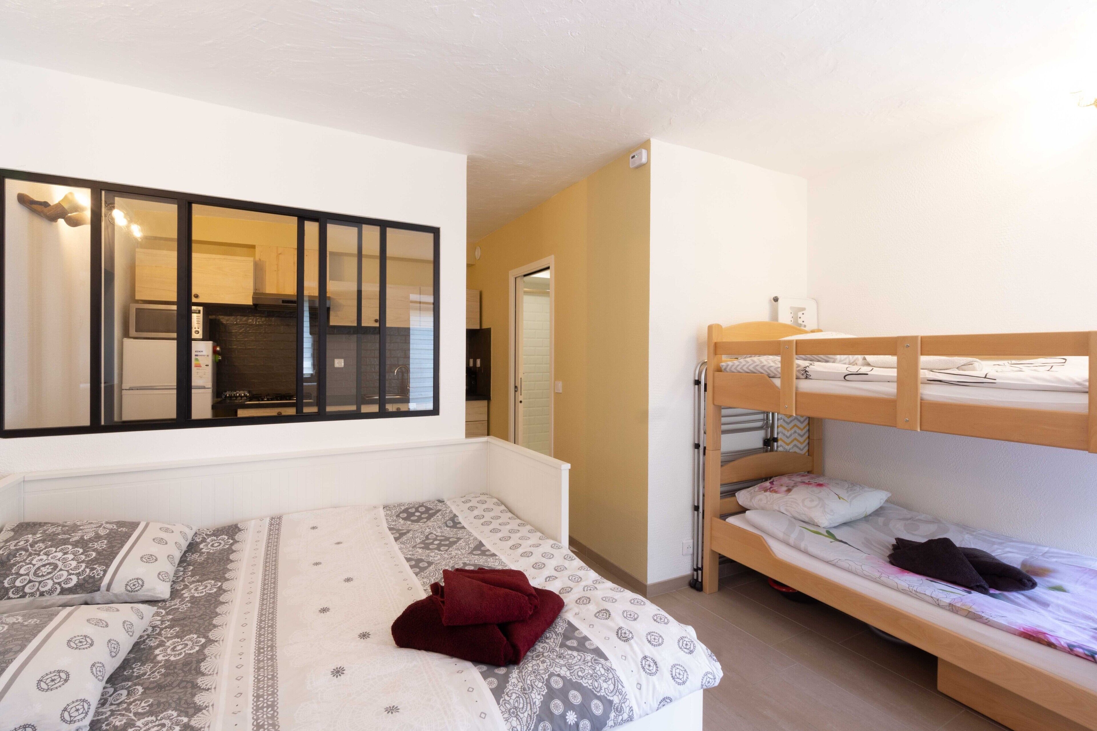 A room with a bunk bed, a single bed, and a kitchenette in the background.