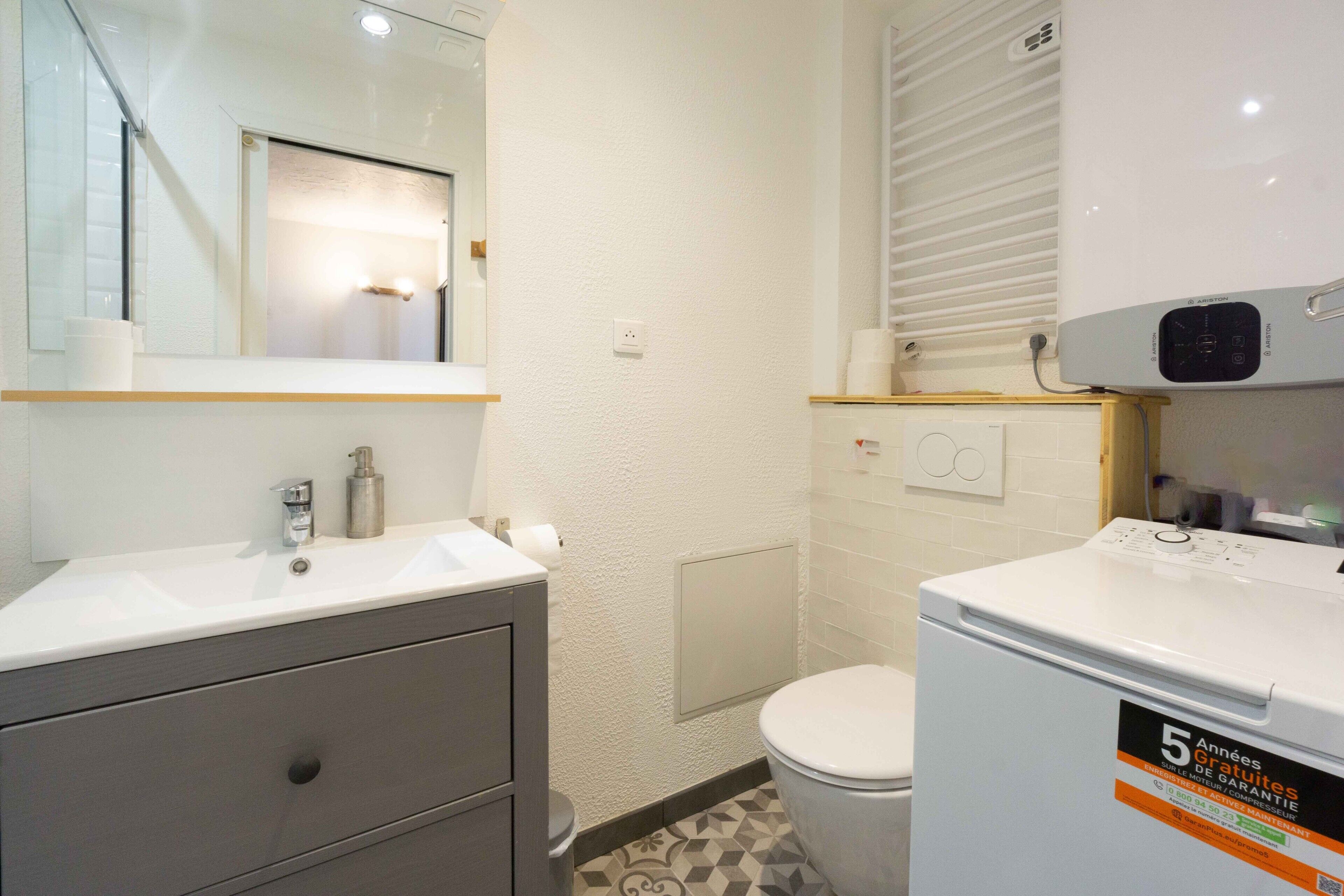 Een badkamer met een witte wastafel, spiegel en toilet. Er hangt een witte verwarming aan de muur en een klein plankje met toiletartikelen.