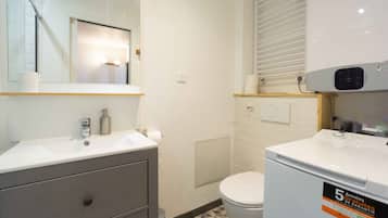 Een badkamer met een witte wastafel, spiegel en toilet. Er hangt een witte verwarming aan de muur en een klein plankje met toiletartikelen.