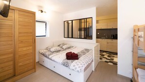 Een slaapkamer met een bed, een houten garderobe, een witte nachtkastje, een spiegel en een keukenhoek met een zwart-wit geruite vloer.
