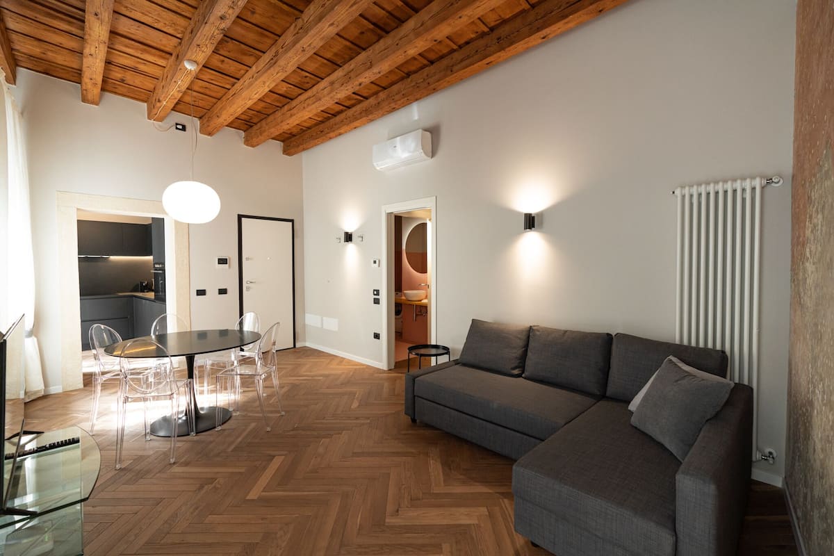 Een moderne woonkamer met een houten plafond, een grijze bank, een eettafel met stoelen en een keuken in de achtergrond.
