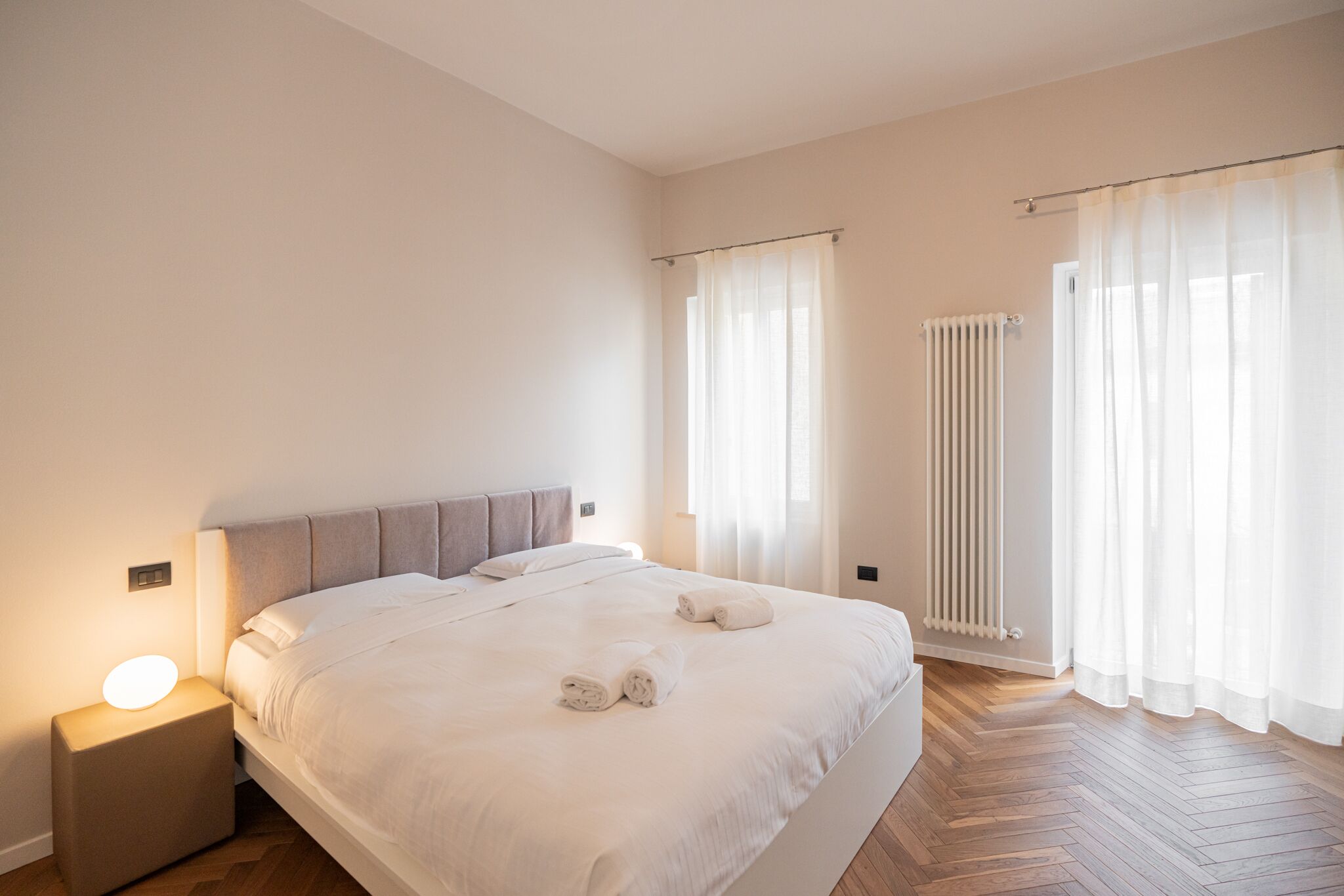 Ein modernes Hotelzimmer mit einem großen Bett, einem Nachttisch, einem Heizkörper und transparenten Vorhängen.