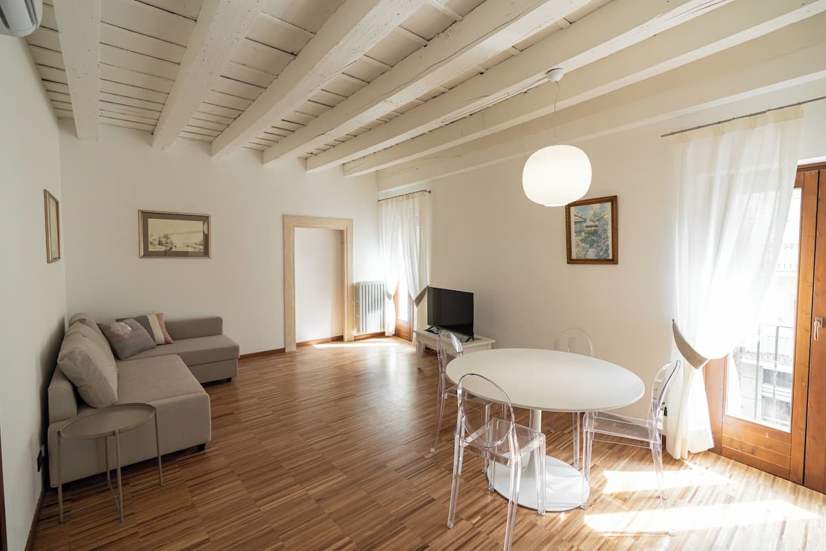 Een moderne woonkamer met een houten vloer, een beige hoekbank, een ronde eettafel met transparante stoelen, een flatscreen televisie en een groot raam met dunne gordijnen.