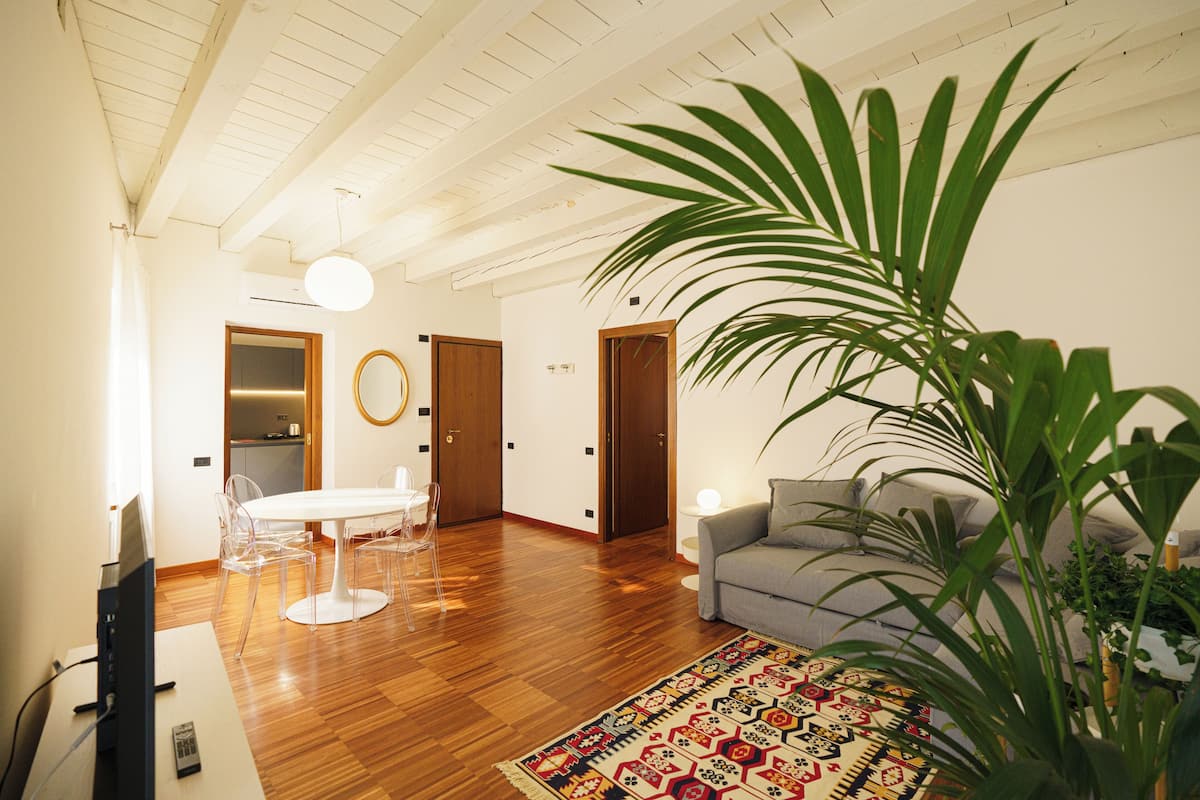 Een ruime woonkamer met een houten vloer, een grijze bank, een eettafel met stoelen en een grote potplant.