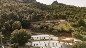 A white building with multiple windows and a flat roof, surrounded by dense forest and a rocky hill in the background. - Holiday Home "Casa Jerzu S'essia de su santu" with Sea View, Garden & Wi-Fi (Jerzu)
