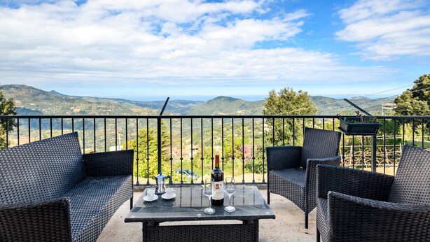 A balcony with wicker furniture, a table with wine glasses and a bottle, and a scenic mountain view. - Holiday Home "Casa Jerzu S'essia de su santu" with Sea View, Garden & Wi-Fi (Jerzu)