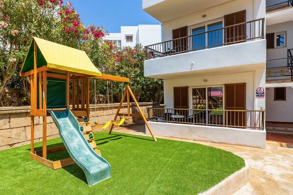 A children's play area with a slide and swing set, surrounded by a well-maintained lawn and a white building with balconies.