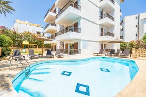 A swimming pool with lounge chairs, a sun umbrella, and a multi-story building with balconies.