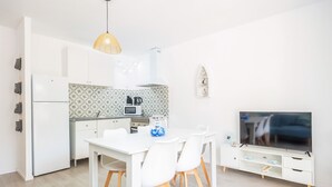Une cuisine moderne avec des meubles blancs, une table à manger avec des chaises, un téléviseur posé sur un meuble blanc et un luminaire suspendu.