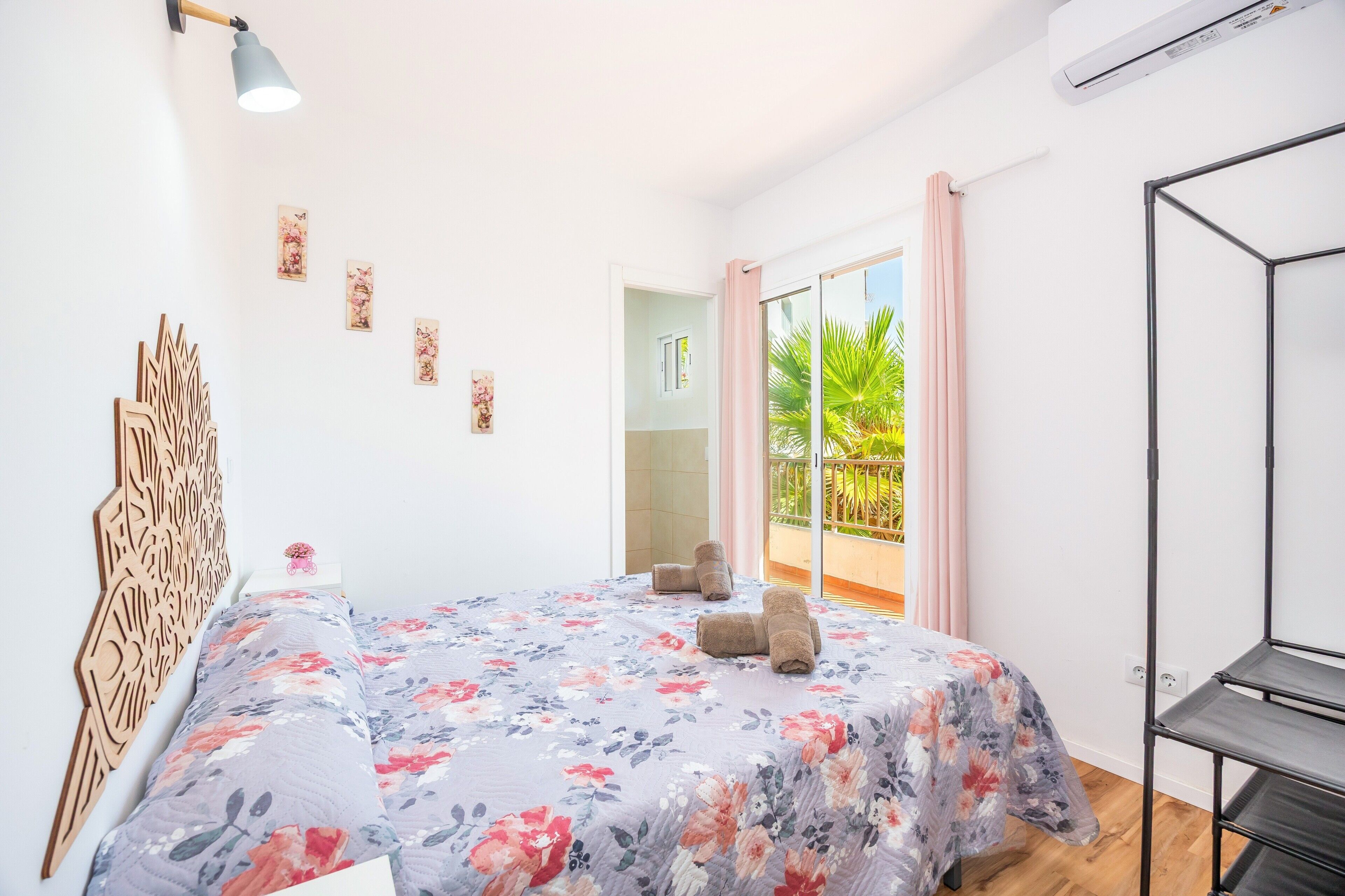 Un dormitorio con cama, cabecera de madera, estante en la pared, ventana con vista a follaje y balcón con cortina rosa.
