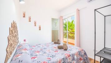 Un dormitorio con cama, cabecera de madera, estante en la pared, ventana con vista a follaje y balcón con cortina rosa.