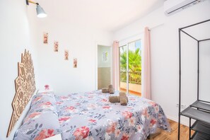 Une chambre avec un lit, une tête de lit en bois, une étagère fixée au mur, une fenêtre donnant sur un paysage verdoyant et un balcon avec un rideau rose.