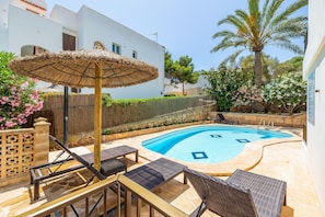 Une piscine entourée de chaises longues et dotée d’un parasol en chaume, le tout bordé d’un bâtiment blanc et de verdure.