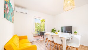 Una habitación luminosa con un sofá amarillo, una mesa de comedor blanca con sillas, un televisor y un balcón con vistas a zonas verdes.