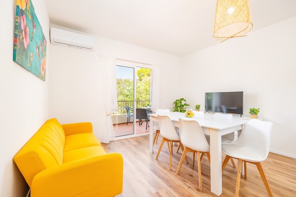 Une pièce lumineuse avec un canapé jaune, une table à manger blanche avec des chaises, un téléviseur et un balcon donnant sur un paysage verdoyant.
