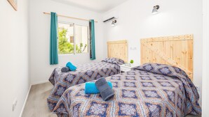 Quarto com duas camas, janela com cortinas, cabeceira de madeira, mesa de cabeceira e luminária fixada na parede.