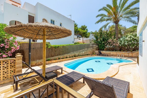 A swimming pool with lounge chairs and a thatched umbrella, surrounded by a white building and greenery.