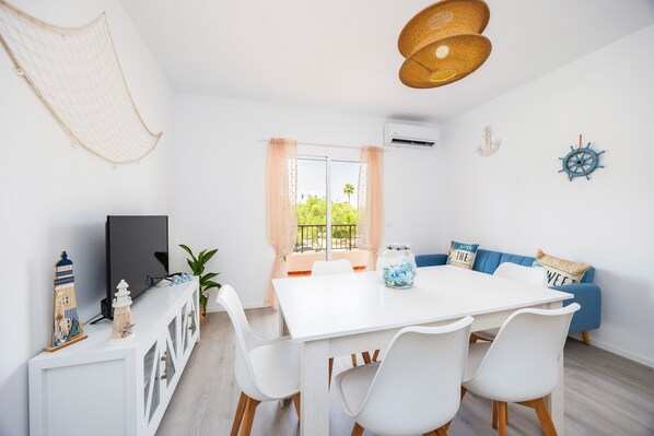 A dining area with a white table, white chairs, a TV, and a window with curtains.