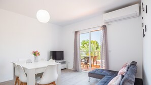 Een moderne woonkamer met een witte eettafel, een bank, een televisie en een balkon met uitzicht op groen.