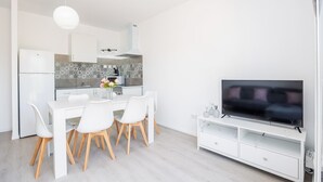 Een moderne keuken met witte kasten, een eethoek met een witte tafel en stoelen, en een flatscreen televisie op een witte standaard.