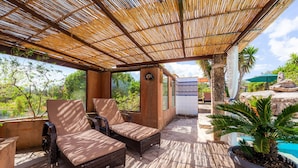 Un espacio de estar al aire libre techado, con mobiliario de mimbre, una planta en maceta y vistas a una piscina y follaje.