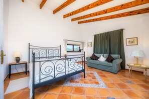 Een slaapkamer met een metalen bedframe, een groene bank, een raam met gordijnen en een betegelde vloer.