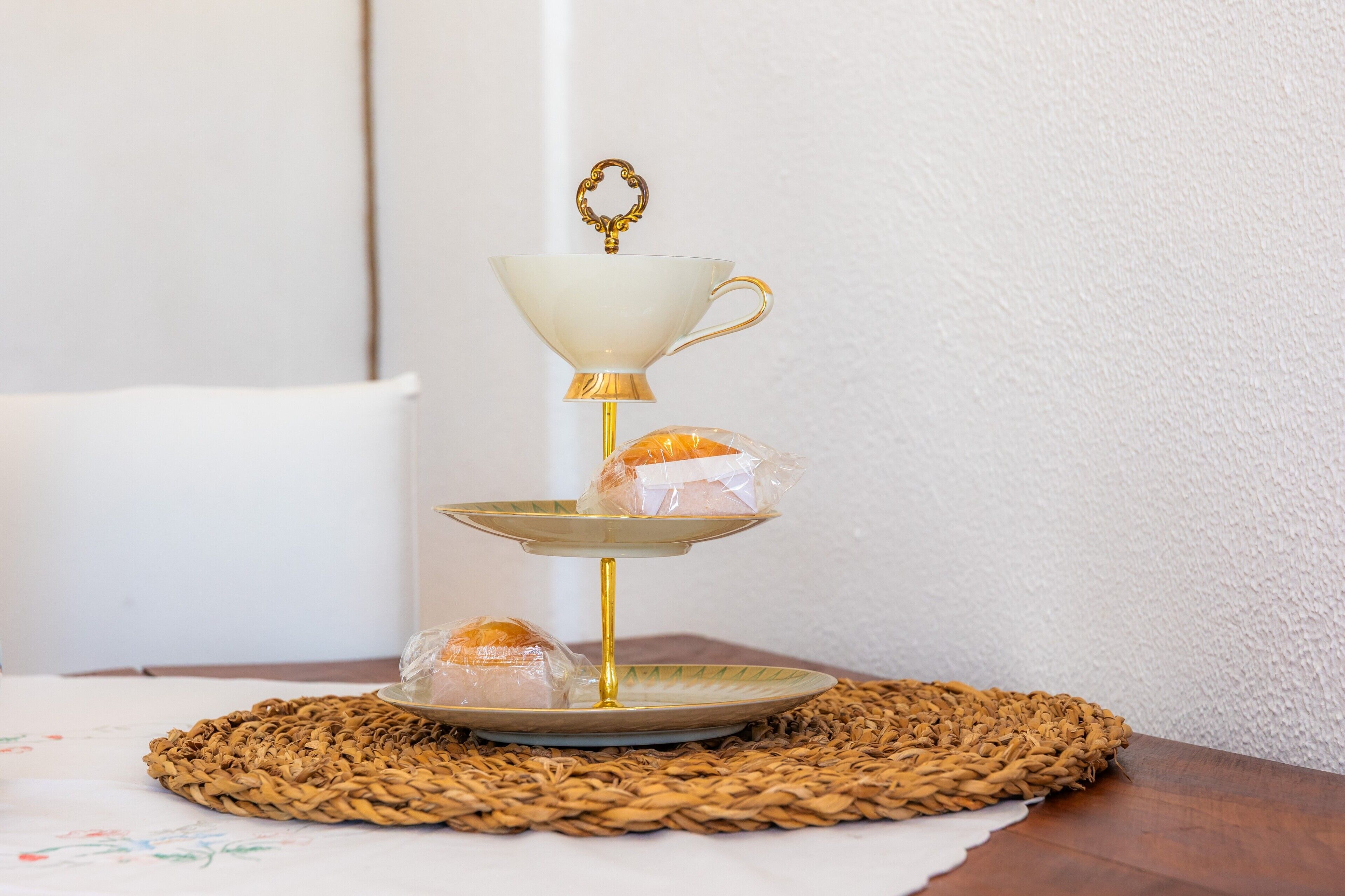 A three-tiered stand with a white cup on top, a plate with a pastry, and a plate with a wrapped item on a woven placemat.