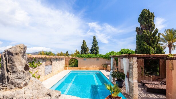 A swimming pool with a stone wall, a lounge area with a sofa, and a view of the sky and trees.