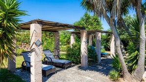 A covered outdoor seating area with lounge chairs, a pergola structure, and palm trees.