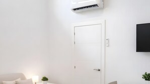 A modern, minimalist room with a sofa, a small table, a dining table with chairs, an air conditioning unit, and a door.