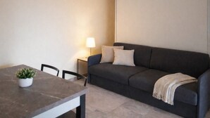 A room with a dark-colored sofa, a small table with a potted plant, a lamp, and a plain wall.