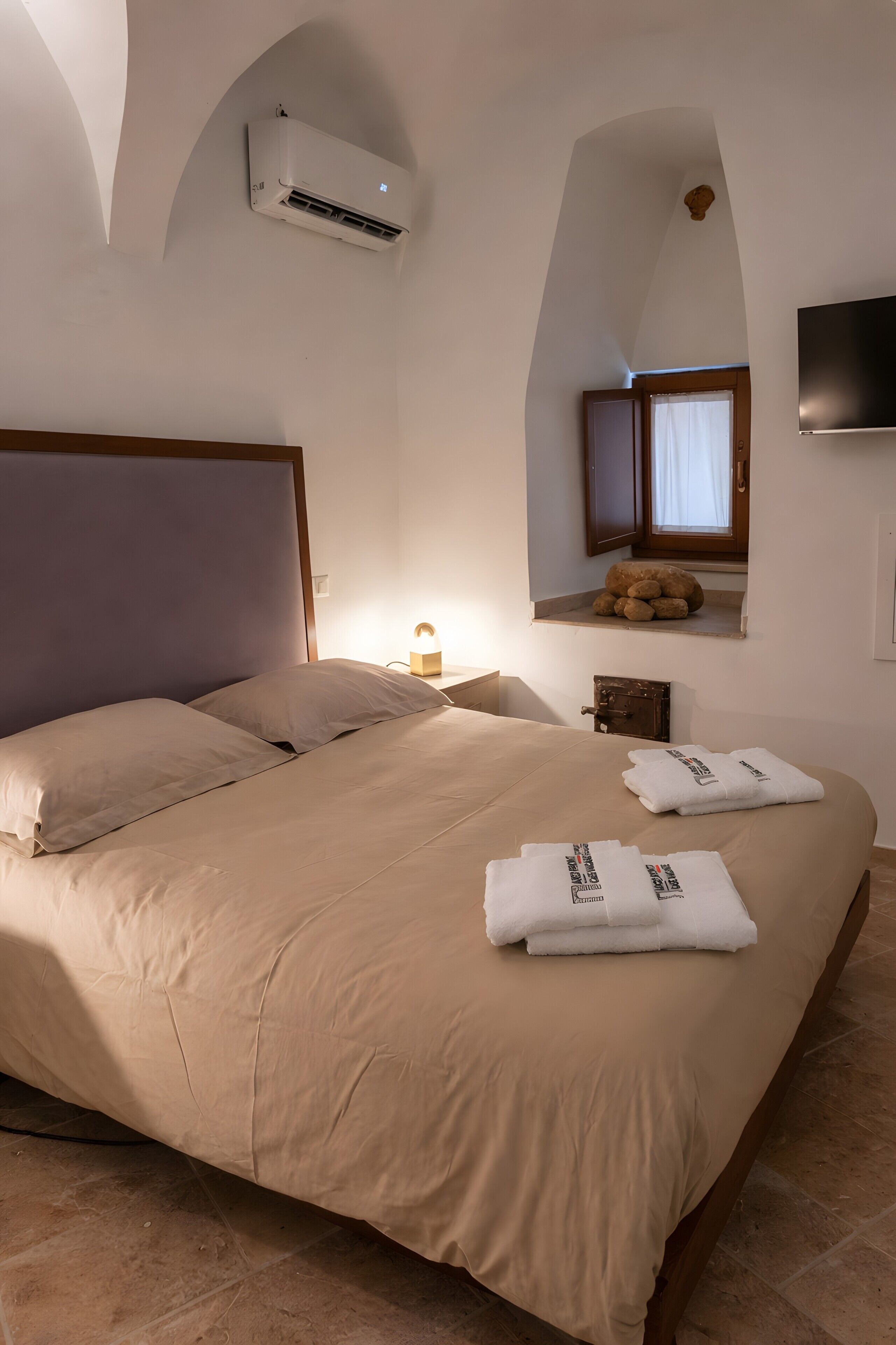 A bedroom with a large bed, an air conditioning unit, a television, a window with a teddy bear, and a bedside lamp.
