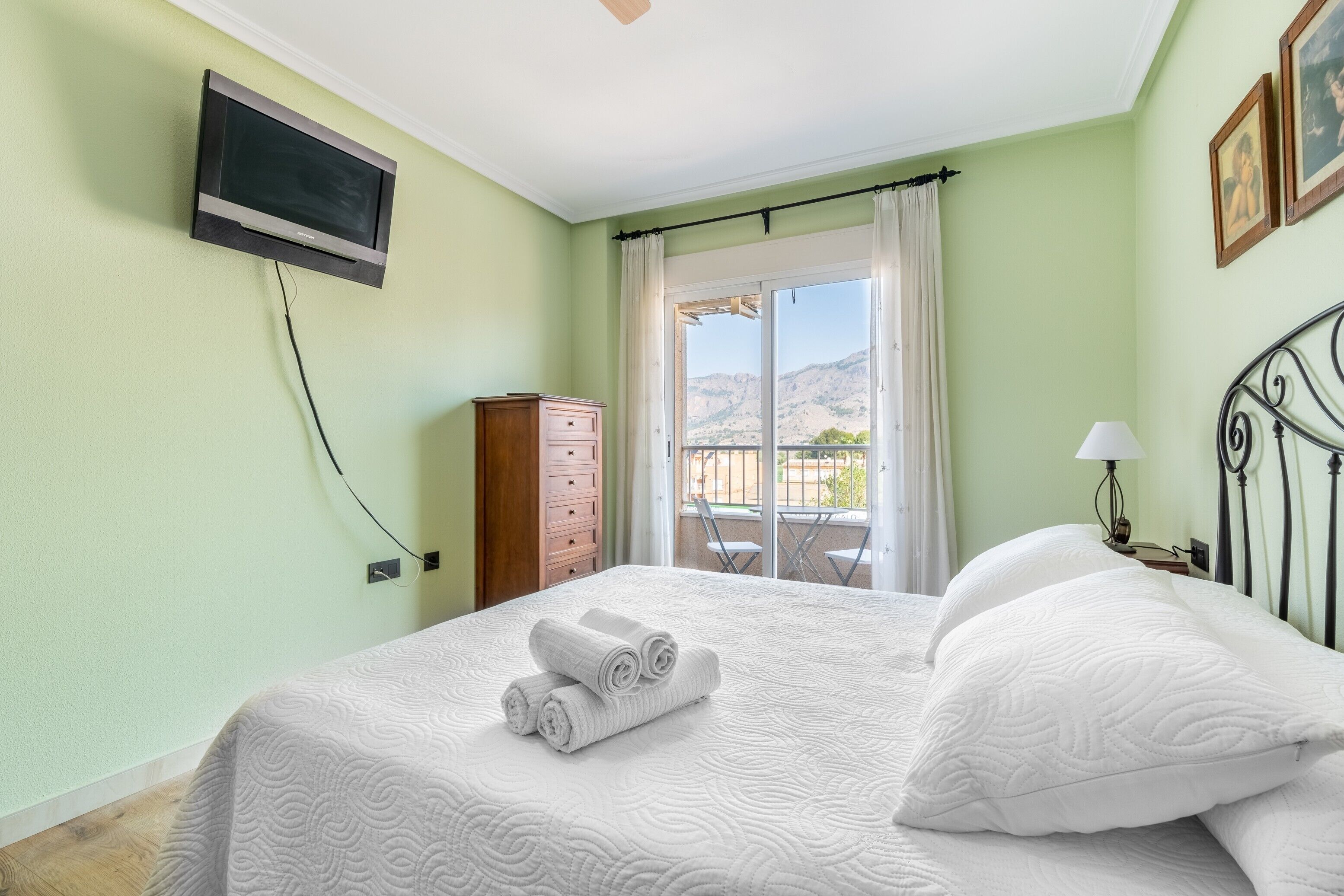 Un dormitorio con cama, televisor montado en la pared, cómoda de madera y balcón con cortinas.