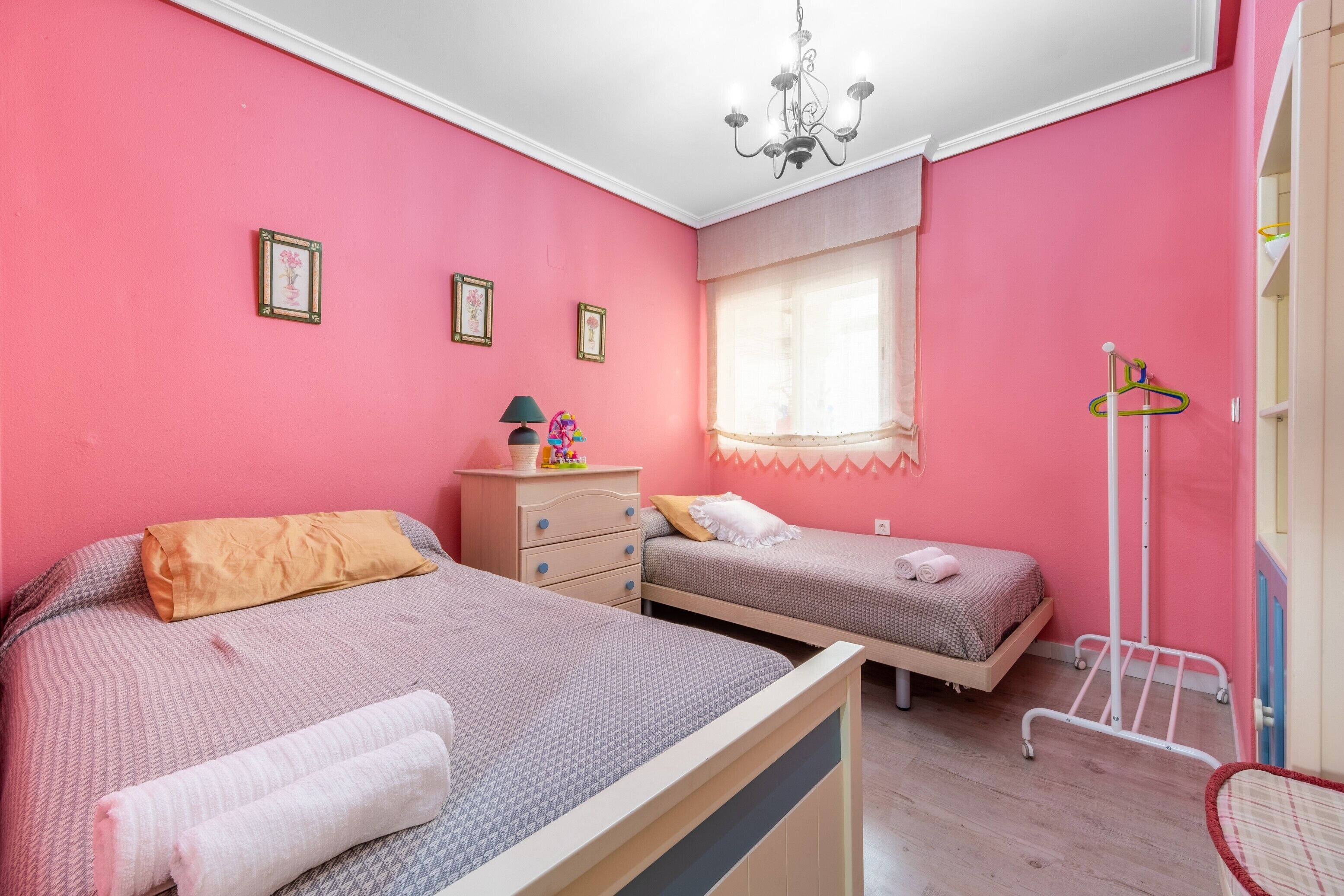 A bedroom with two beds, a dresser, a window with sheer curtains, a hanging light fixture, and a coat rack.