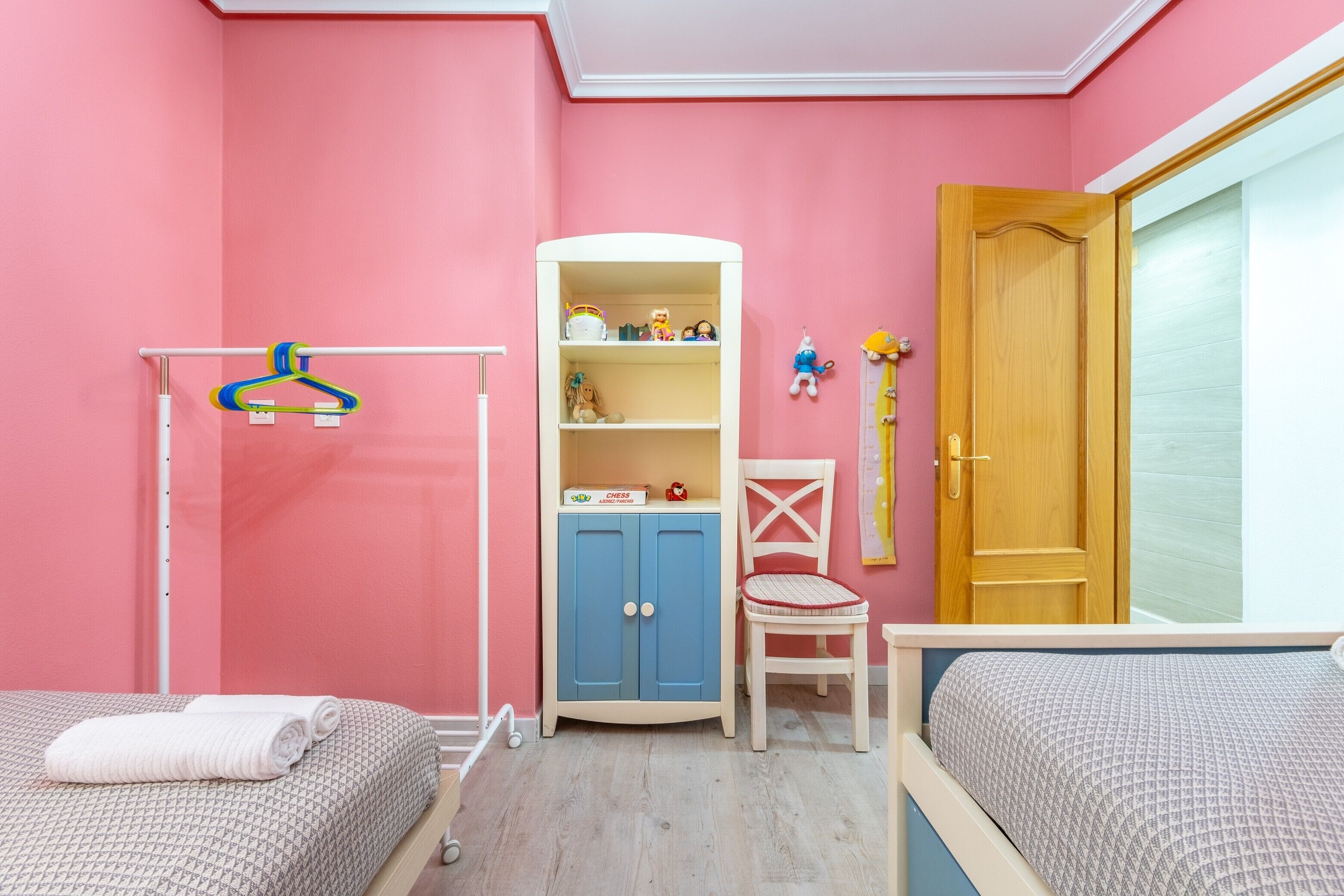 Une pièce aux murs roses, avec un radiateur blanc, une armoire bleue, une porte en bois et deux lits.