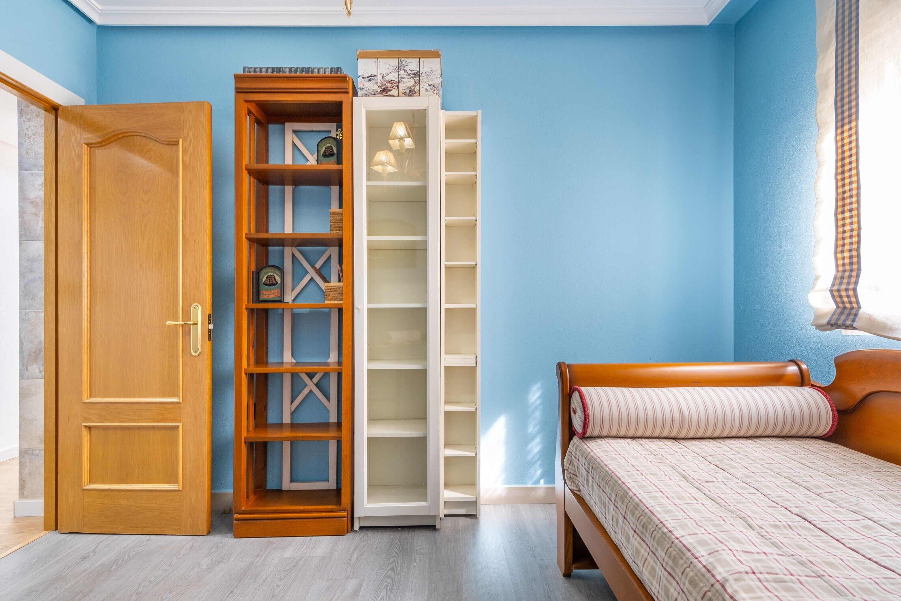 Un dormitorio con puerta de madera, armario con estantes múltiples, cama con colcha estampada y pared azul.