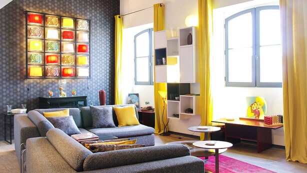 Un salon moderne avec un canapé gris, des coussins jaunes, un luminaire fixé au mur et un coin repas agrémenté de rideaux jaunes.