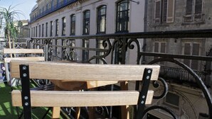 En træbænk med metalstøtter foran en bygning med flere vinduer og en balkon.