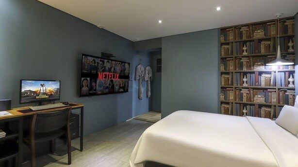 Een slaapkamer met een bed, een bureau, een televisie, een boekenplank en een wand met een poster.