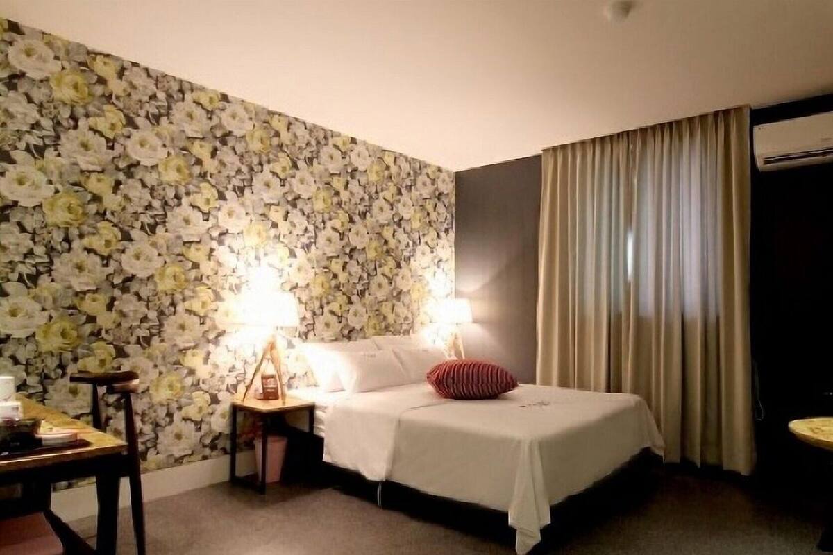 Une chambre d’hôtel avec un papier peint à motifs floraux, un lit avec une literie blanche, une table de chevet avec une lampe et une fenêtre avec des rideaux.