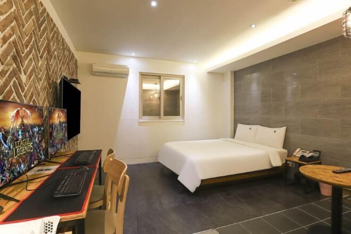 Habitación de hotel con cama, escritorio con computadora, silla, mesita y televisor montado en la pared.