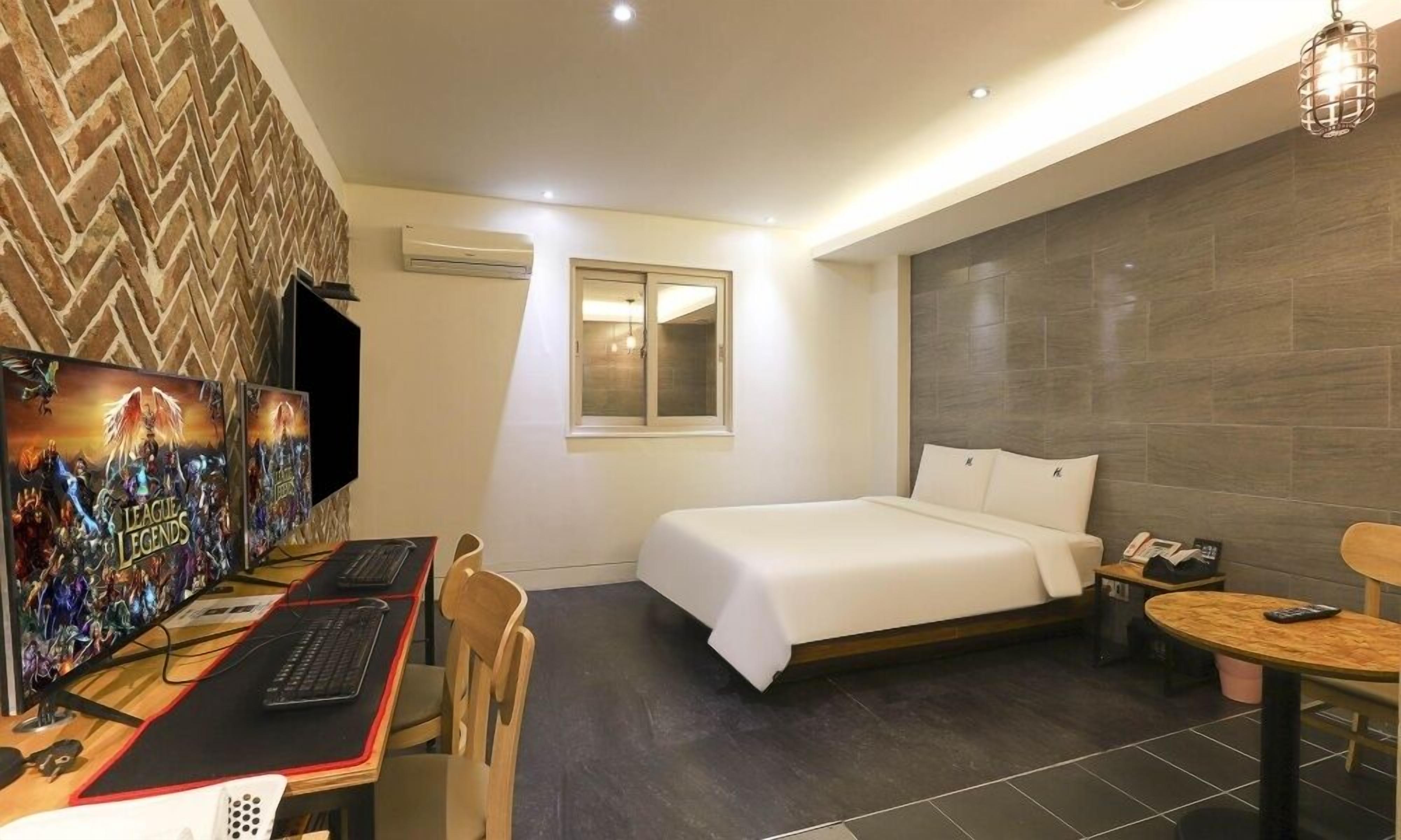 Habitación de hotel con cama, escritorio con computadora, una silla, una mesita y televisor mostrando 'League of Legends'.